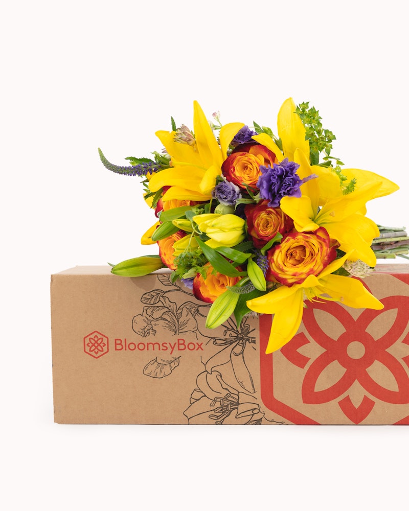 A vibrant bouquet of yellow lilies, orange roses, and purple flowers protruding from a BloomsyBox cardboard box against a white background.