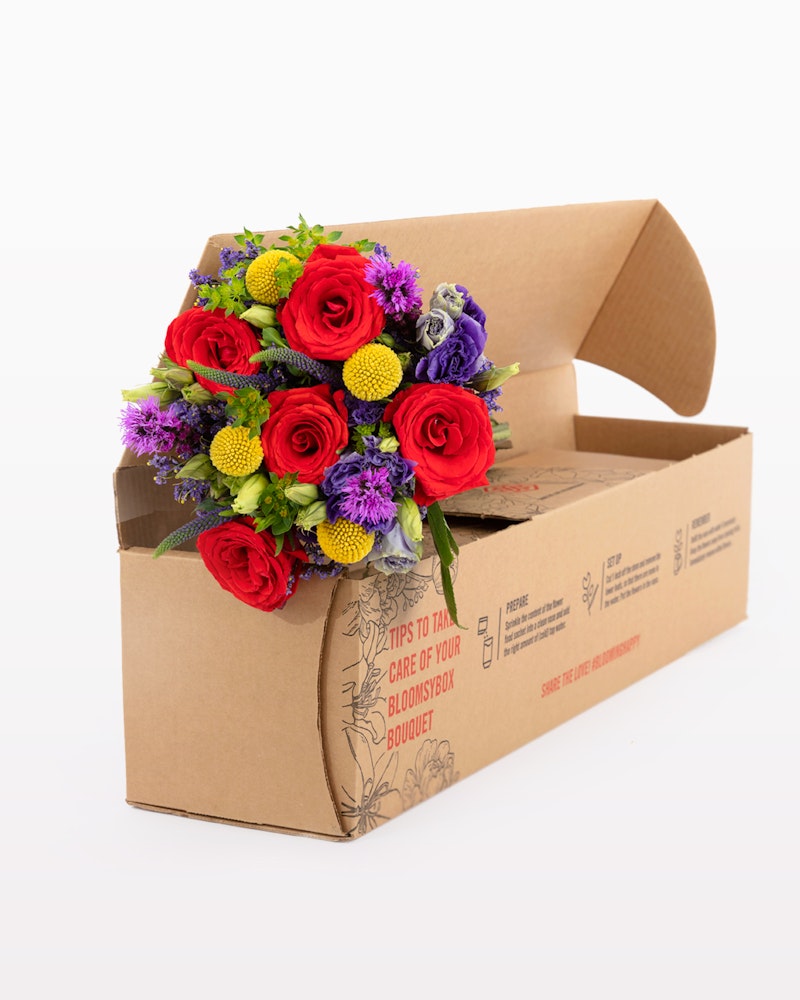 Vibrant bouquet of red roses, purple flowers, and yellow blooms packaged neatly in a brown cardboard box with care instructions for delivery.