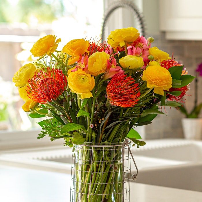 Vibrant bouquet of yellow roses, red-orange pincushion proteas, and pink flowers in a glass vase on a kitchen counter with a window in the background.