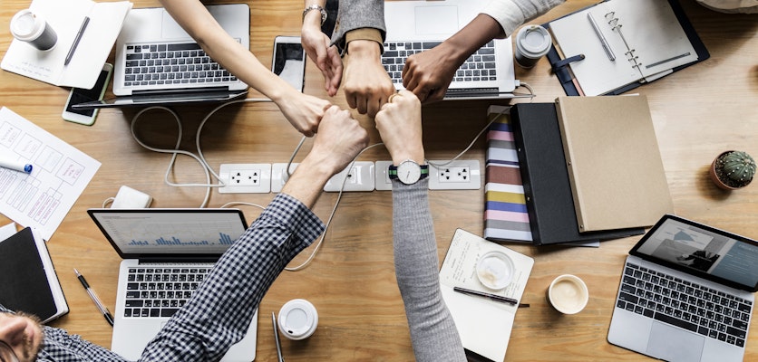 Collaborative workspace with diverse hands giving a fist bump over laptops and notebooks.