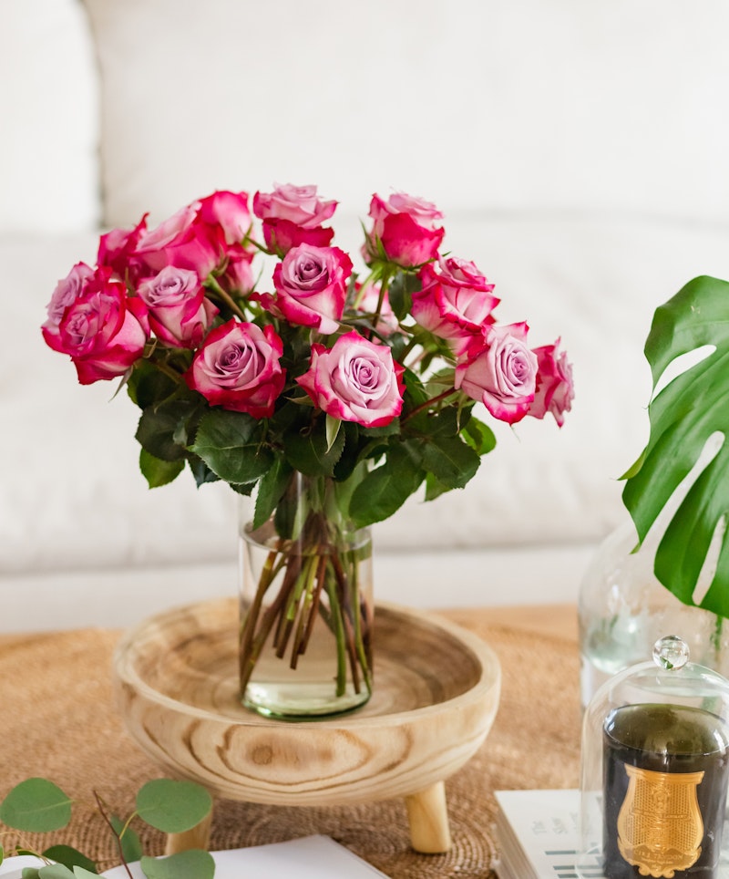 Bouquet of vibrant pink roses arranged in a clear glass vase on a wooden tray with a comfortable white sofa and green leafy plant in the background.