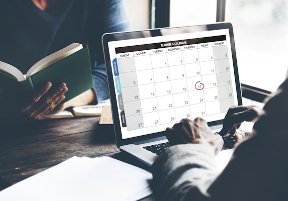 A person planning their schedule with a laptop and calendar, surrounded by books.