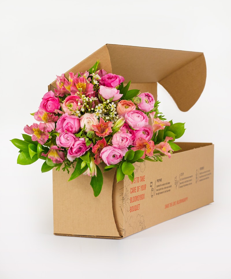 Vibrant bouquet of pink roses and assorted flowers emerging from a cardboard box against a white background, symbolizing fresh flower delivery services.