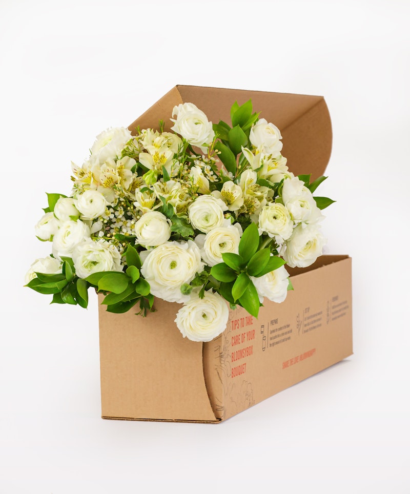 Beautiful white roses and green foliage arranged elegantly in a cardboard flower box with a visible postal label, showcased against a clean, white background.
