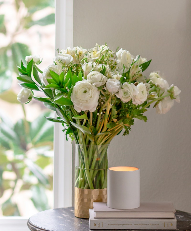 Elegant white flowers arranged in a clear vase on a wooden table beside a lit candle, with natural light casting a warm glow, near a window with garden view.