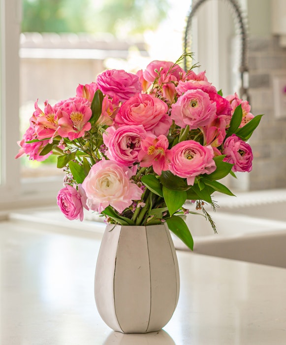 Vibrant bouquet of pink flowers including roses and peonies in a white vase on a table with natural light streaming in from a window in the background.