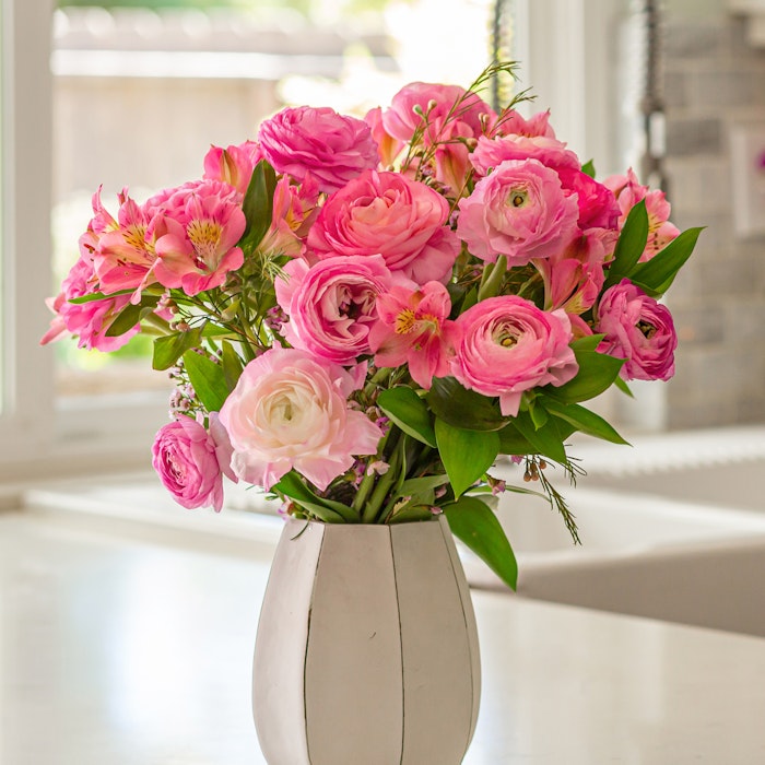 Vibrant bouquet of pink flowers including roses and peonies in a white vase on a table with natural light streaming in from a window in the background.