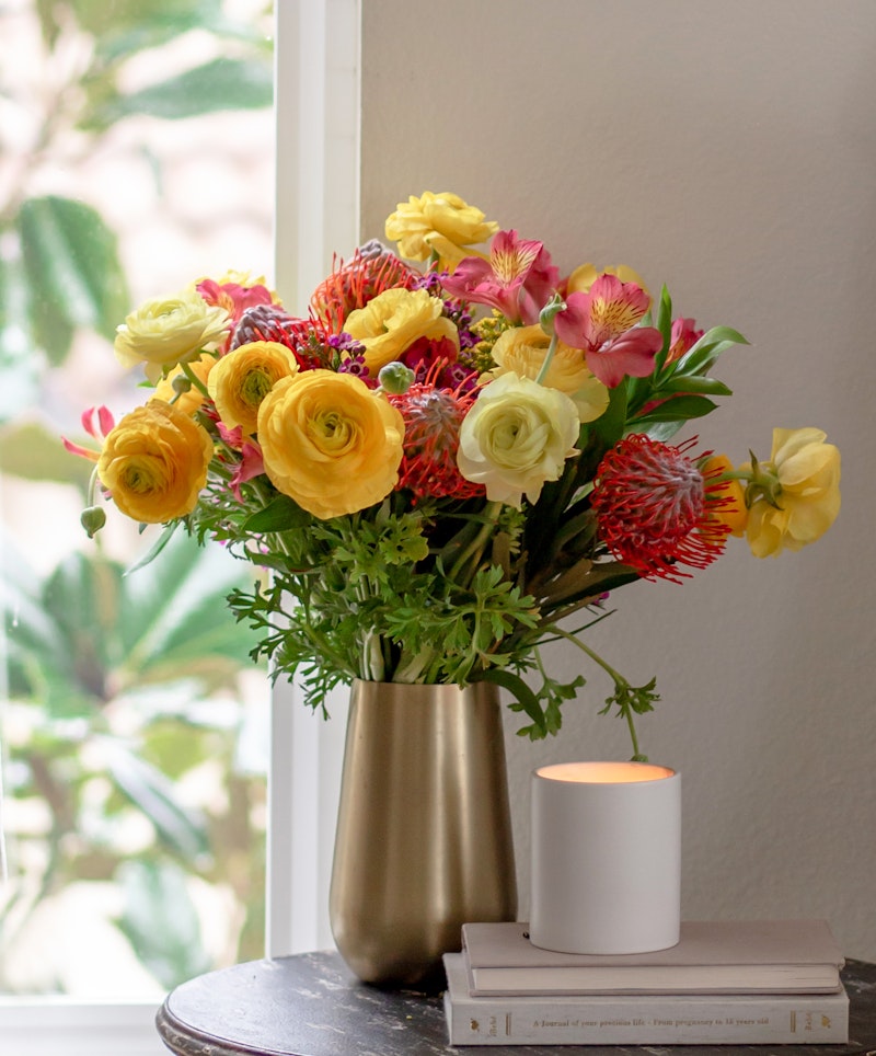 A vibrant bouquet of yellow and pink flowers in a gold vase on a table with books beside a lit candle, in a cozy room with natural light.