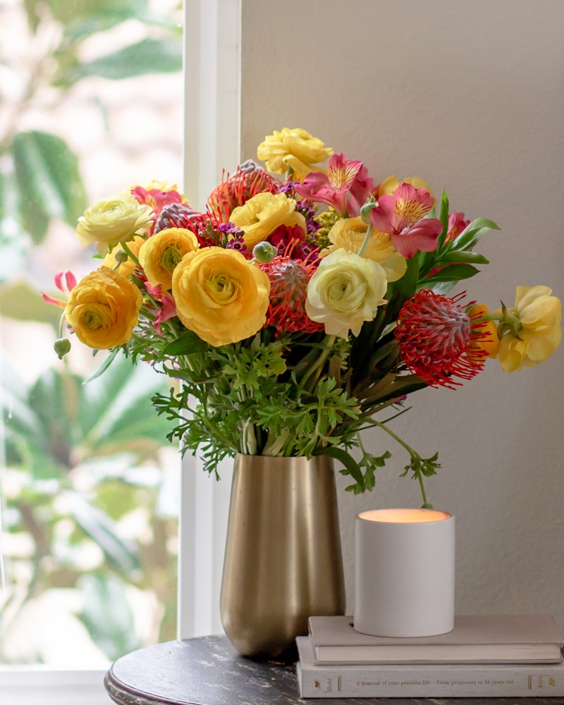 A vibrant bouquet of yellow and pink flowers in a gold vase on a table with books beside a lit candle, in a cozy room with natural light.