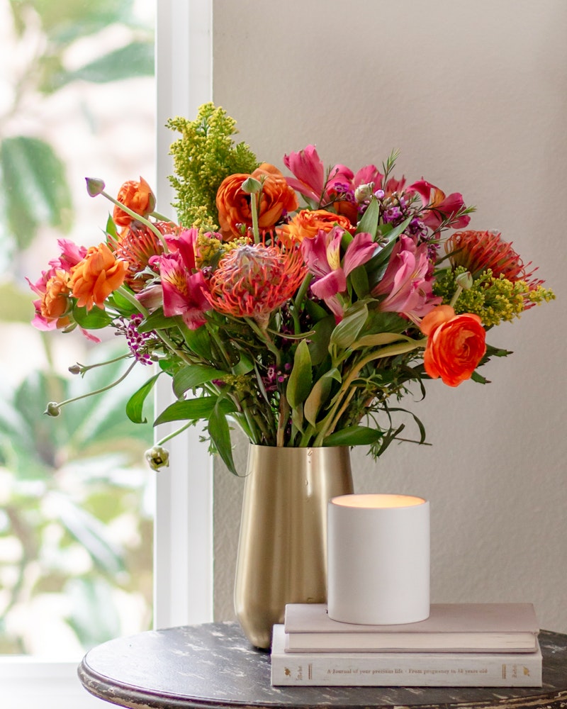 Bouquet of vibrant orange and pink flowers in a golden vase on a table with books and a lit candle, by a window with natural light.