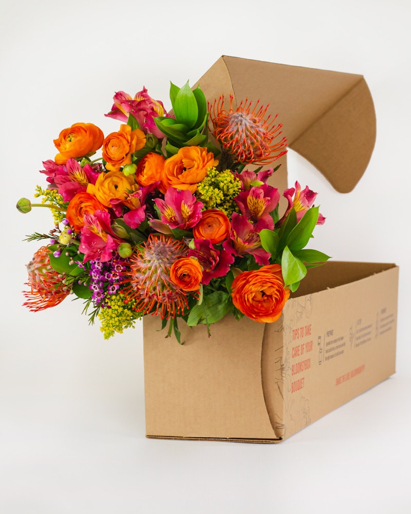 Vibrant bouquet of orange roses, purple flowers, and lush greenery spilling from an open cardboard box against a white background, conveying a fresh delivery.