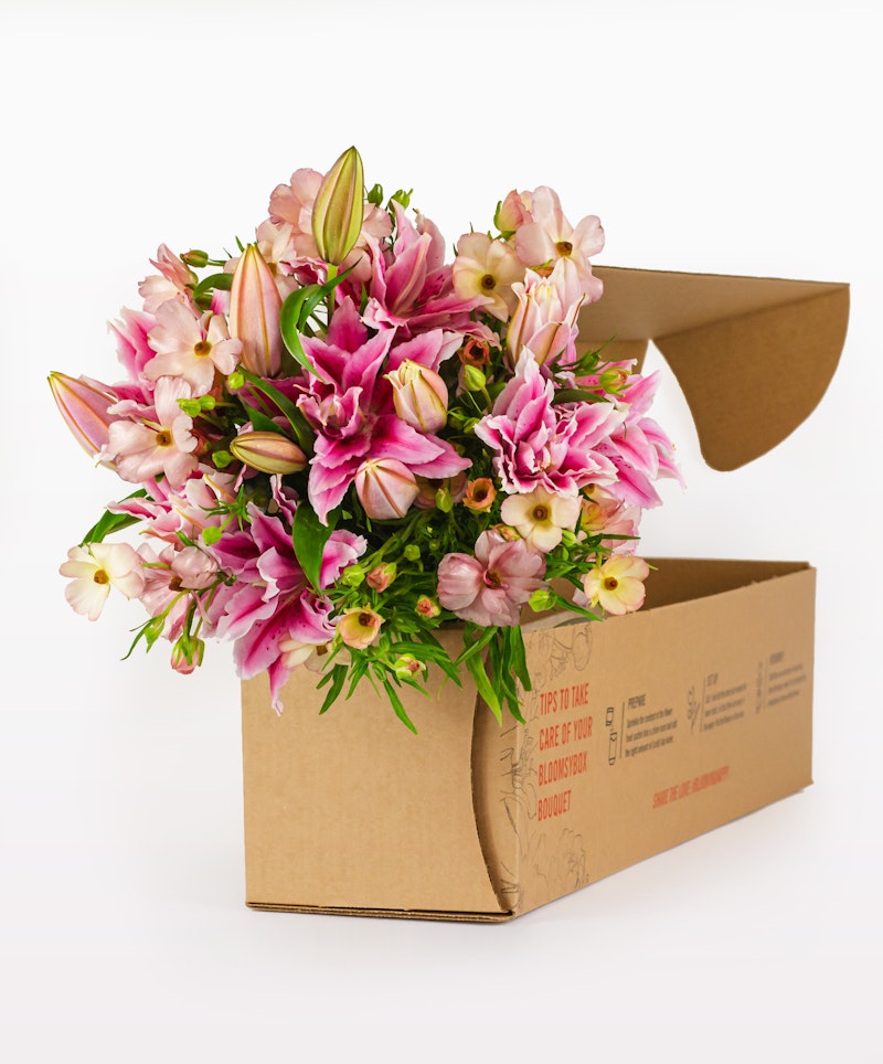 A vibrant bouquet of pink lilies and other flowers emerging from a brown cardboard delivery box against a white background, ready for gifting.