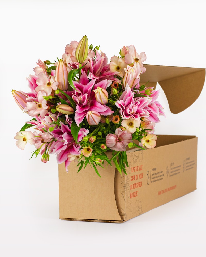 A vibrant bouquet of pink lilies and other flowers emerging from a brown cardboard delivery box against a white background, ready for gifting.