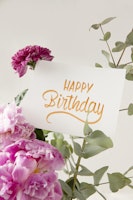Elegant birthday card with gold script surrounded by a bouquet of pink and purple flowers and greenery against a neutral background, conveying warm birthday wishes.