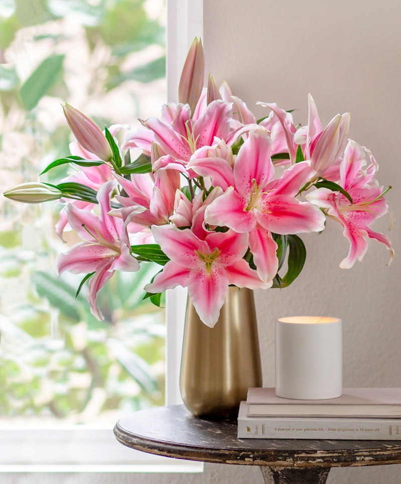 Vibrant pink lilies arranged in a gold vase on a wooden table beside stacked books and a lit candle, with a blurred garden view through a window in the background.