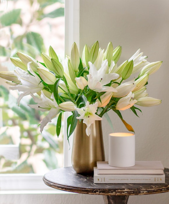 A bouquet of fresh white lilies in a golden vase on a round table beside a glowing candle and stacked white books, near a window with green foliage outside.