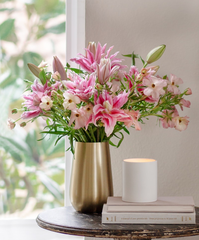 A bouquet of pink lilies and other flowers in a gold vase on a table beside a white candle and books, with a lush green plant visible through the window.