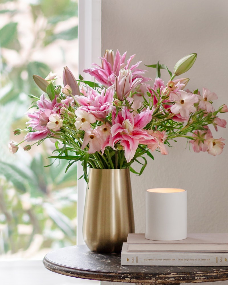 A bouquet of pink lilies and other flowers in a gold vase on a table beside a white candle and books, with a lush green plant visible through the window.
