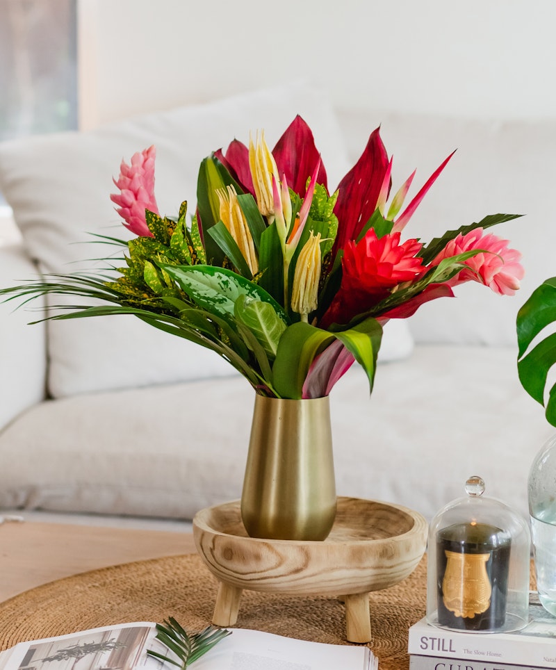 Vibrant tropical flowers in a gold vase on a wooden table, with a white sofa, coffee table books, and decorative items in a cozy living room setting.