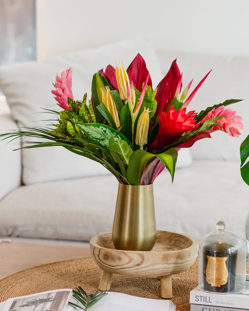 Vibrant tropical flowers in a gold vase on a wooden table, with a white sofa, coffee table books, and decorative items in a cozy living room setting.