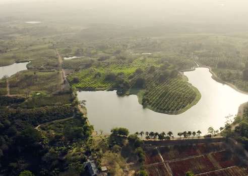 Aerial view of a serene lake surrounded by lush greenery and agricultural fields, with a glimpse of a rural landscape and winding paths under a hazy sky.
