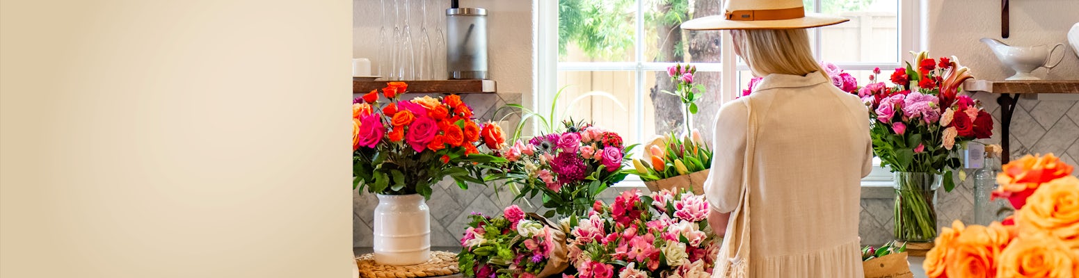 A woman in a sun hat admires vibrant floral arrangements in a bright, cheerful shop.