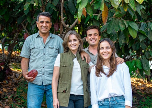 A family of four smiling in a cacao farm, with the man holding a cacao pod, surrounded by lush greenery, depicting a healthy outdoor lifestyle and agriculture.
