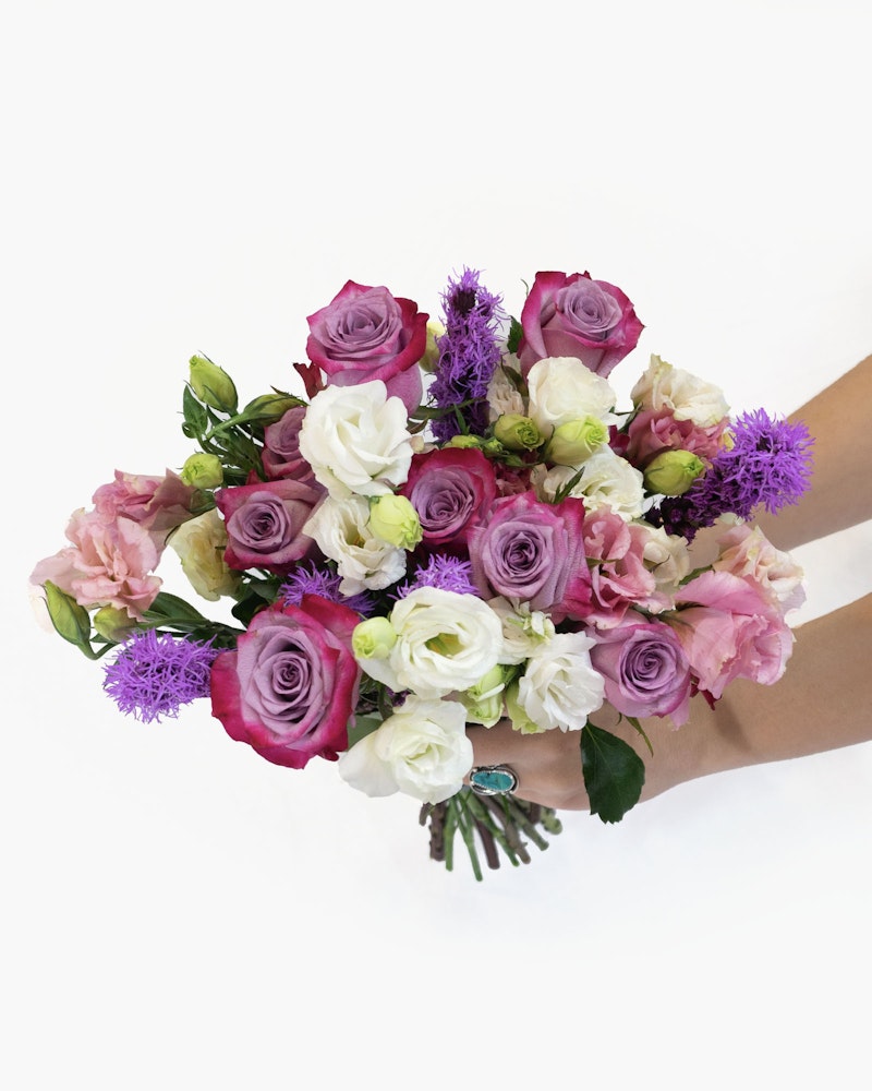 A person holding a vibrant bouquet of flowers featuring pink and purple roses, white blooms, and greenery, set against a white background.