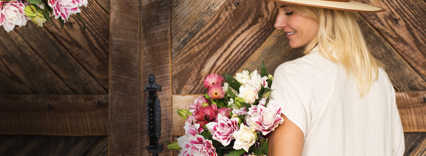A woman in a wide-brimmed hat holding a vibrant floral bouquet against a rustic backdrop.