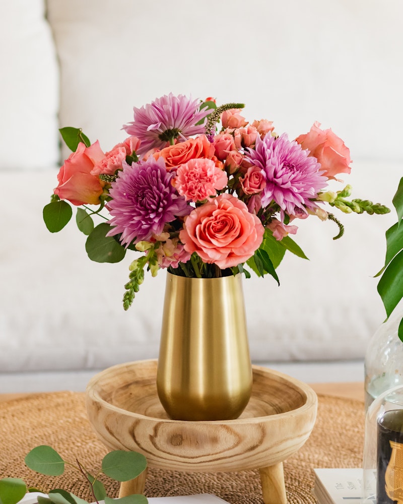 Vibrant bouquet of fresh flowers featuring pink roses, purple dahlia, and complementary foliage in a gold vase on a wooden tray, with a cozy white sofa in the background.