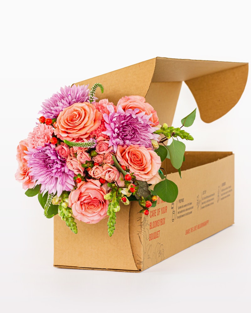 Vibrant bouquet of pink and orange flowers, including roses and dahlias, artistically arranged in a cardboard flower box on a white background.
