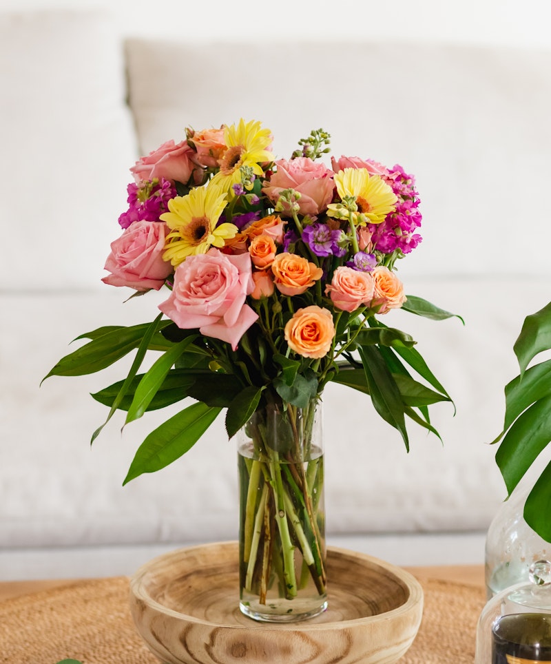 A vibrant bouquet of flowers with pink roses, orange blooms, and yellow lilies in a clear glass vase on a wooden tray against a white couch background.