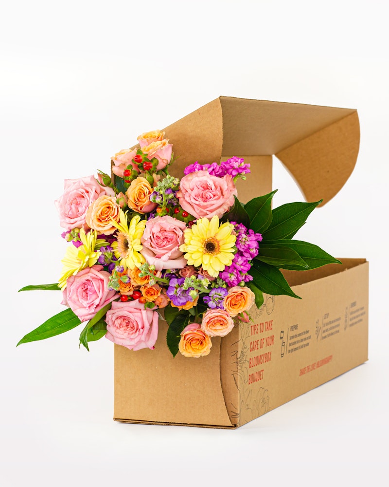 Vibrant bouquet of fresh flowers including pink roses, yellow blooms, and purple accents peeking from a cardboard flower delivery box on a white background.