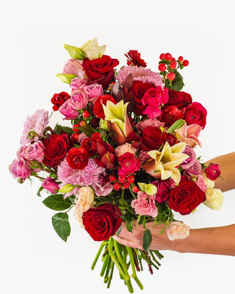 A person's hands holding a lush bouquet of flowers featuring red roses, pink blooms, and yellow lilies against a white background, with a fresh, elegant, and natural look.