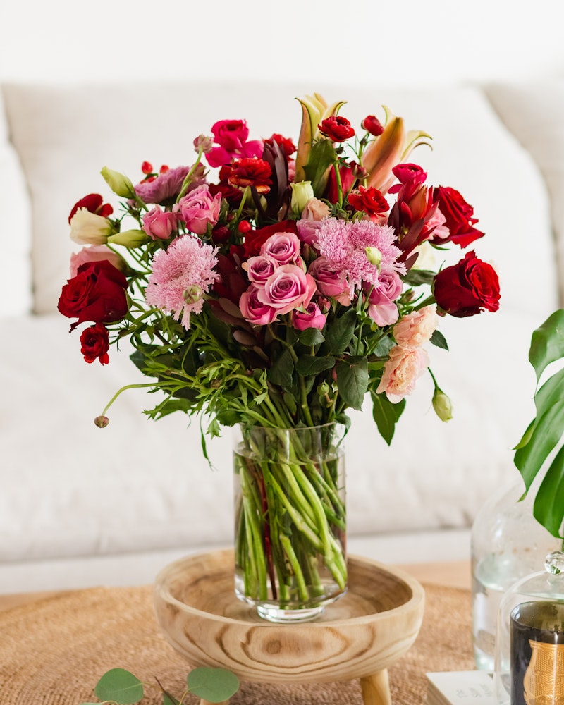 A vibrant bouquet of red roses, pink lilies, and scabiosa flowers with green foliage arranged in a glass vase on a wooden tray, set against a cozy white couch backdrop.