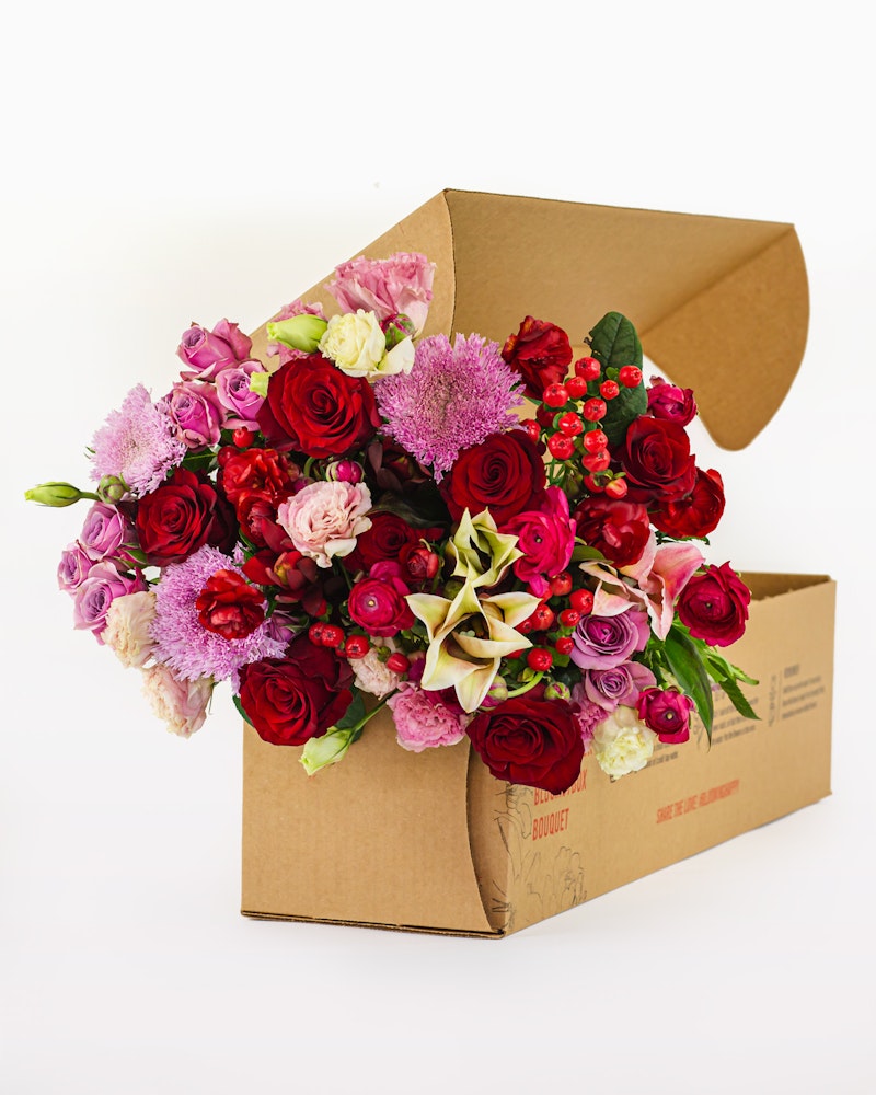 A vibrant bouquet of mixed flowers including roses, lilies, and other blooms overflowing from a cardboard flower delivery box against a white background.