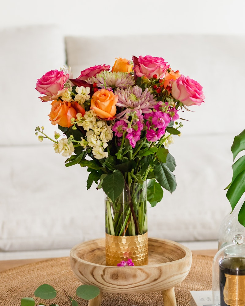 A vibrant bouquet of flowers with pink roses, orange blooms, and purple accents in a gold-trimmed glass vase, set on a wooden tray in a cozy living room setting.