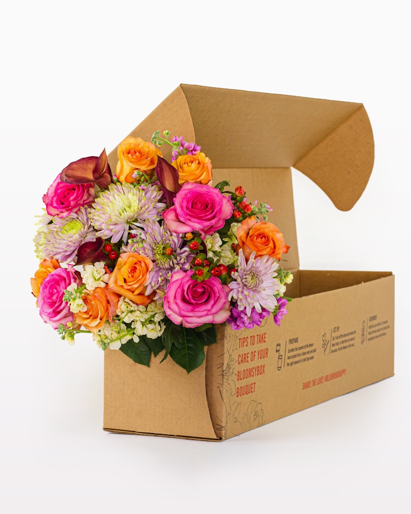 A vibrant bouquet of pink roses, orange blooms, and purple flowers arranged neatly in an open cardboard box against a white background.