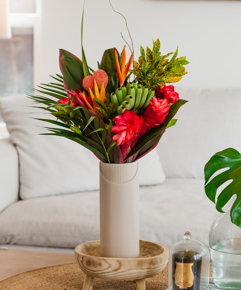 A vibrant bouquet of exotic flowers, including orange and red blooms and green leaves, displayed in a white vase on a wooden stand in a cozy living room setting.