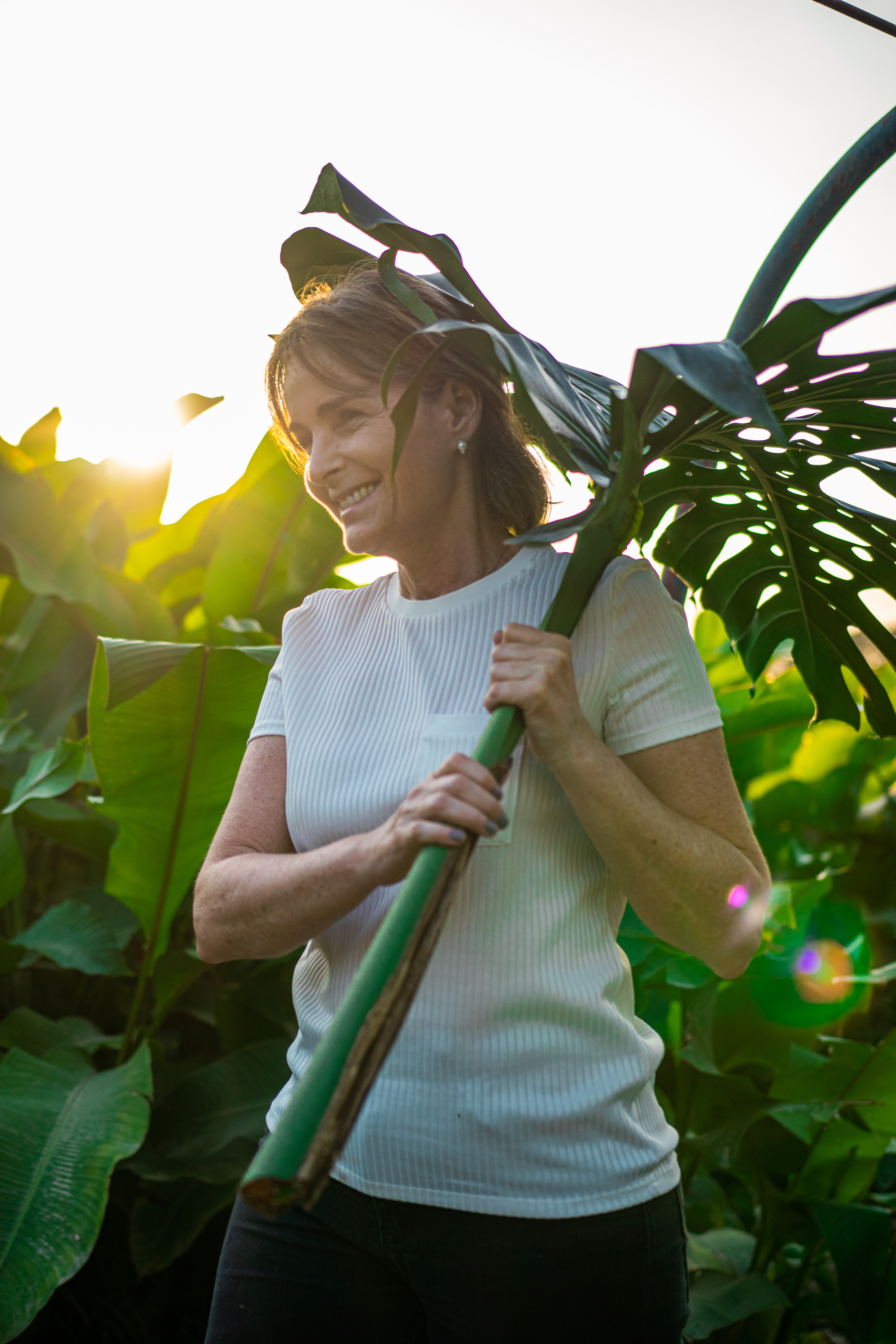 Woman smiling while playfully holding a large green leaf in a lush garden setting.