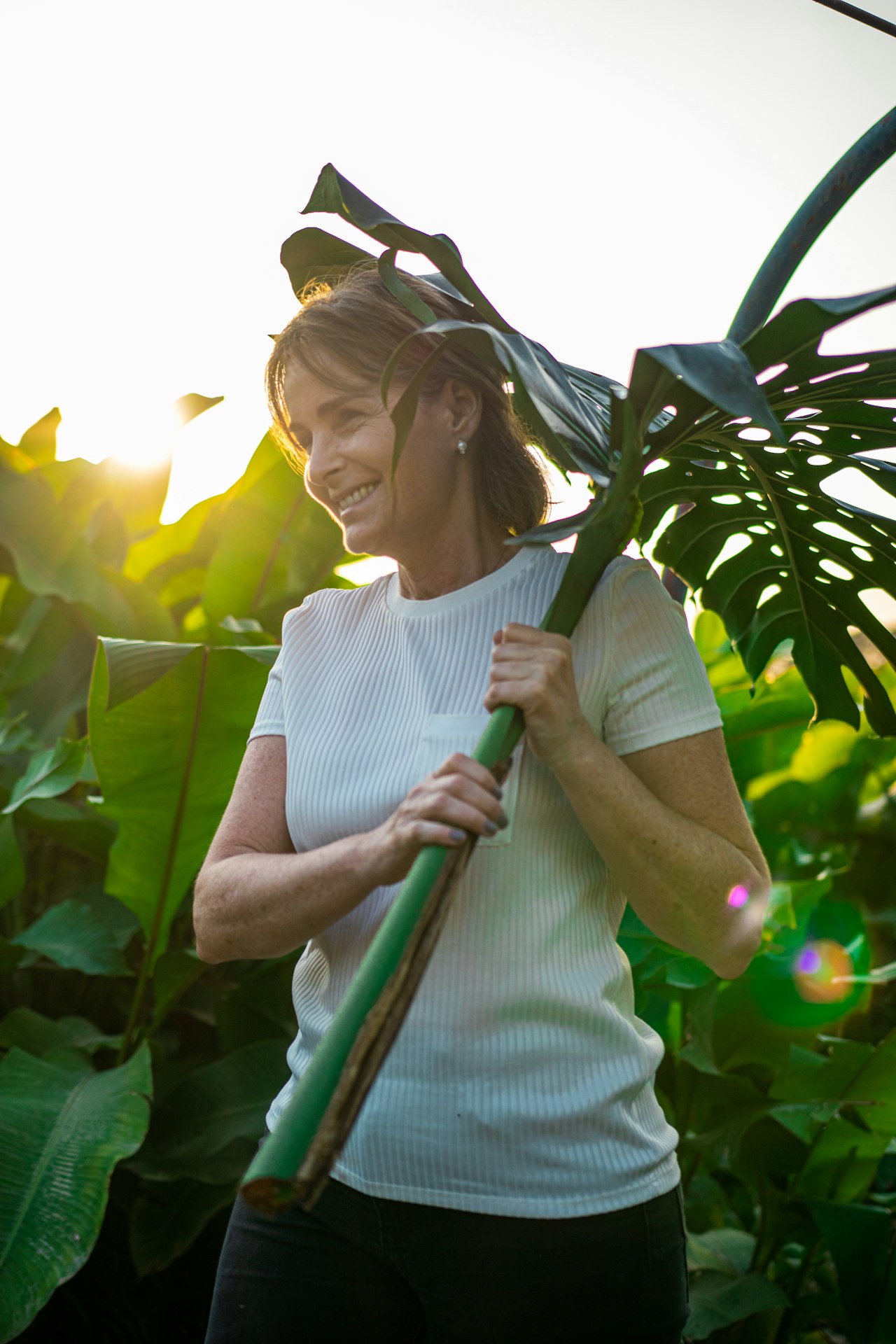 Smiling woman standing amid large green leaves on a sunny day, holding a long-handled gardening tool over her shoulder with sun flares in the background.