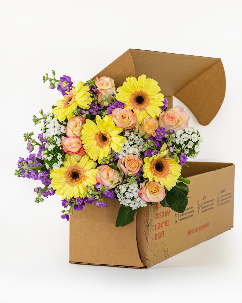 Fresh bouquet of yellow sunflowers, peach roses, and purple accents spilling from a cardboard delivery box on a white background, symbolizing online flower delivery services.