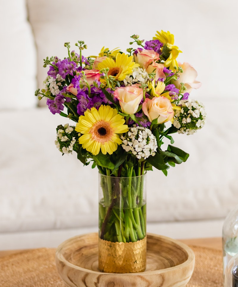 A vibrant bouquet of flowers featuring yellow gerberas, pink roses, purple accents, and greenery arranged in a golden vase set on a wooden tray against a soft backdrop.