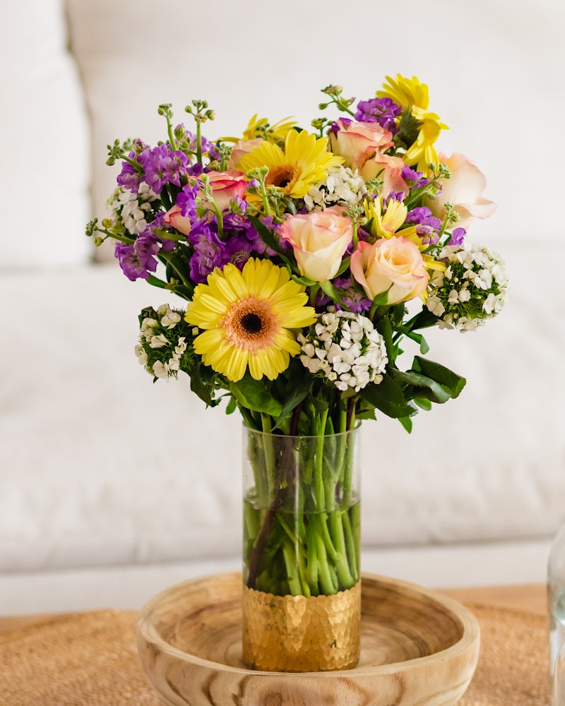 A vibrant bouquet of flowers featuring yellow gerberas, pink roses, purple accents, and greenery arranged in a golden vase set on a wooden tray against a soft backdrop.