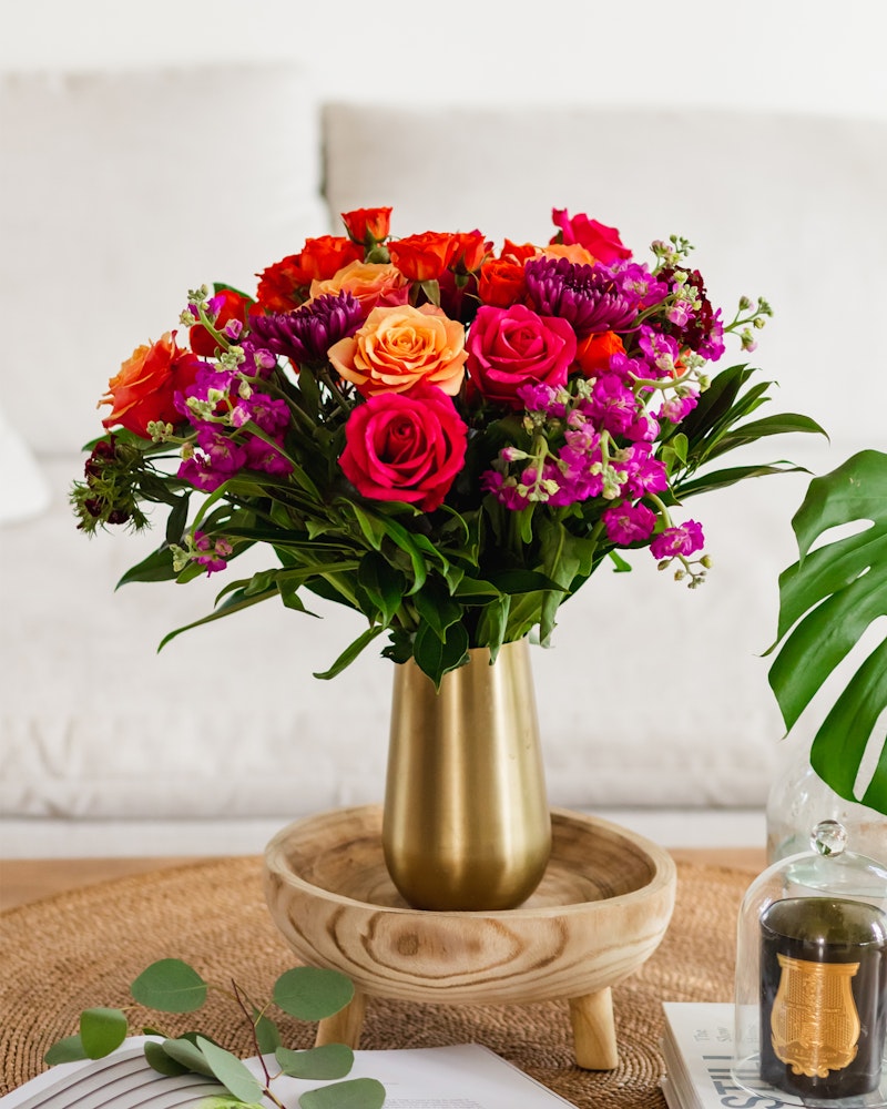 A vibrant bouquet of orange and pink roses, purple flowers, and greenery arranged in a gold vase on a wooden tray, with a tropical leaf and home decor in the background.