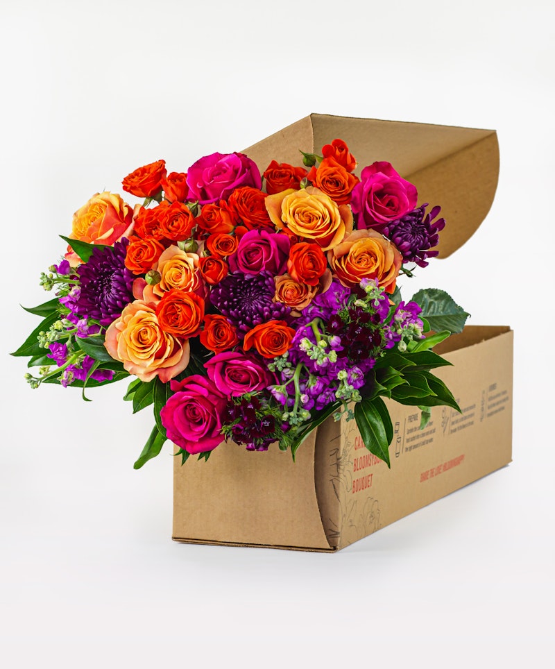 Vibrant bouquet of fresh roses in shades of orange, pink, and purple with lush green foliage, elegantly arranged and partially packaged in a cardboard box on a white background.