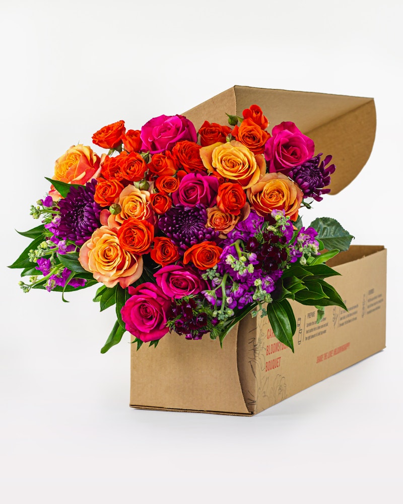 Vibrant bouquet of fresh roses in shades of orange, pink, and purple with lush green foliage, elegantly arranged and partially packaged in a cardboard box on a white background.