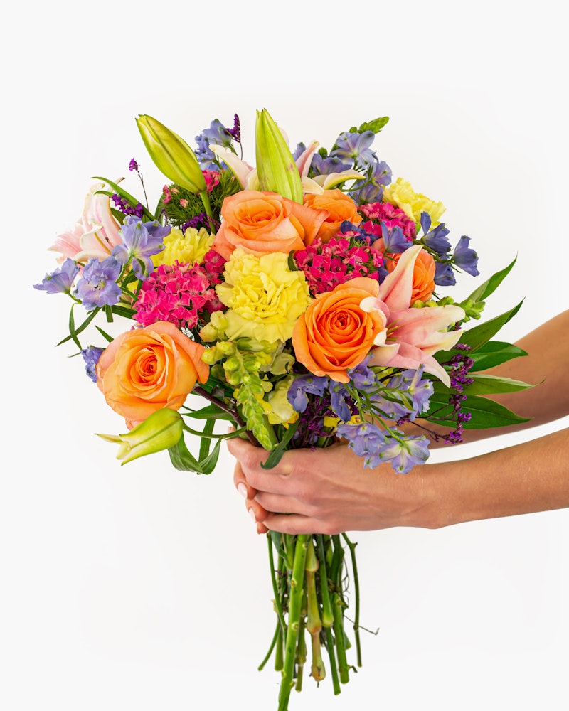Vibrant bouquet of flowers featuring orange roses, purple larkspurs, pink hydrangeas, and yellow carnations held by hands against a white background.