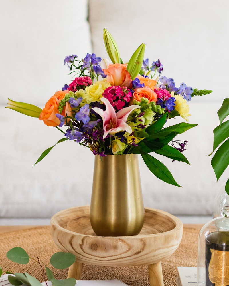 Vibrant bouquet of flowers including pink lilies, orange roses, and purple blooms arranged in a golden vase on a wooden stand with a white textured background.