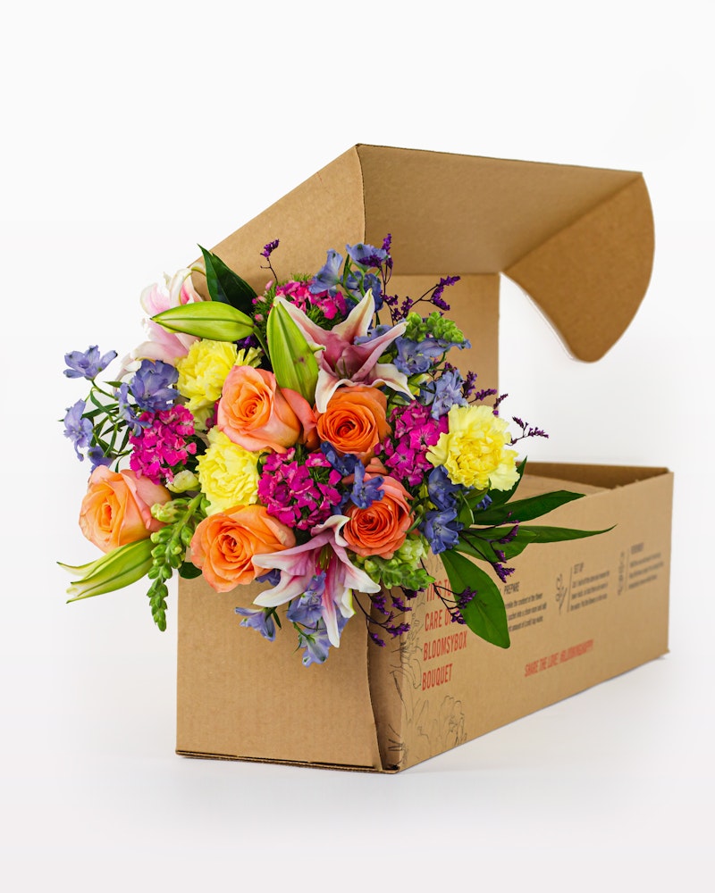 Vibrant bouquet of colorful flowers including orange roses and yellow blooms, artistically arranged in an open cardboard flower box on a white background.